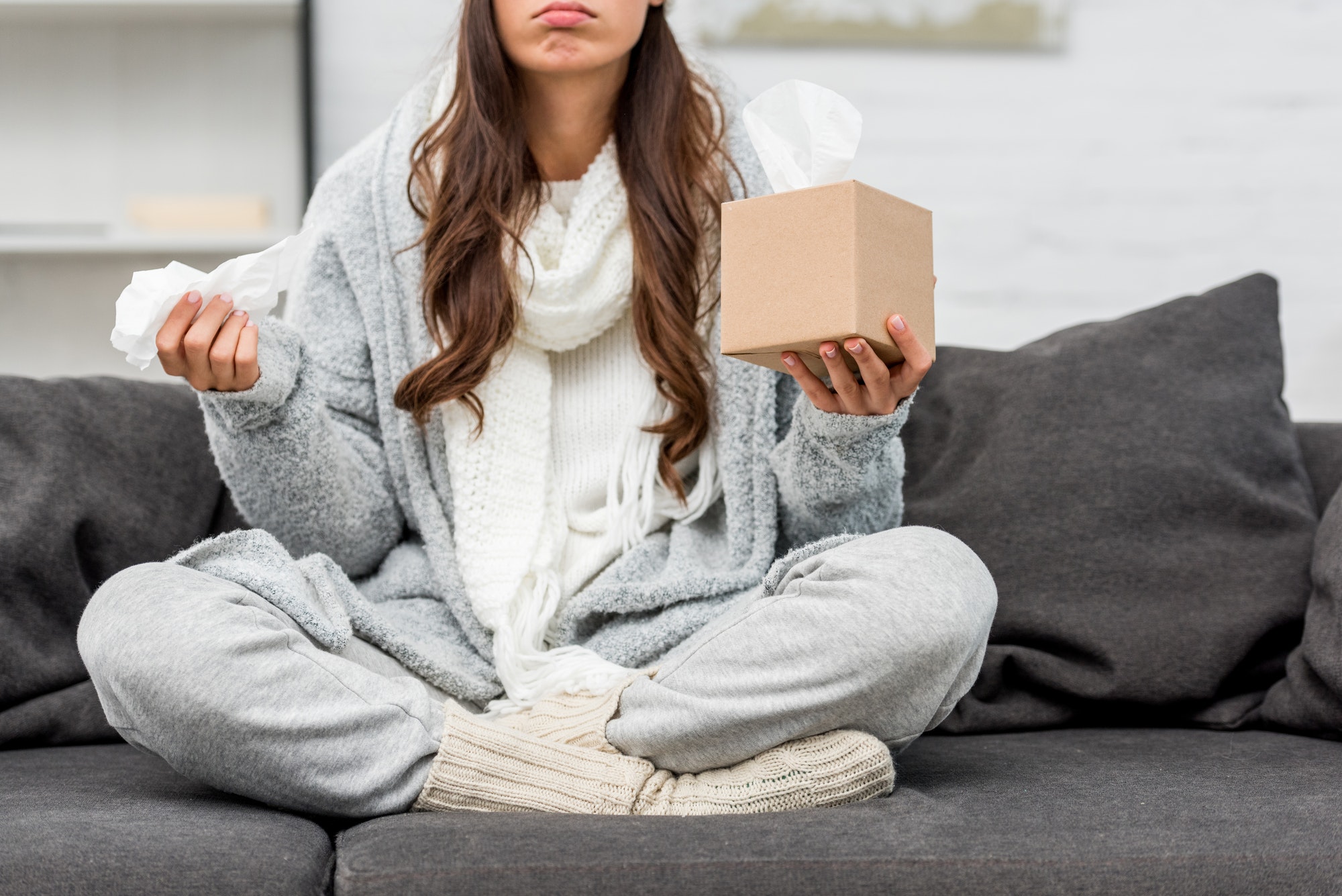 cropped shot of sick young woman in warm clothes sitting on couch at home and holding box of paper